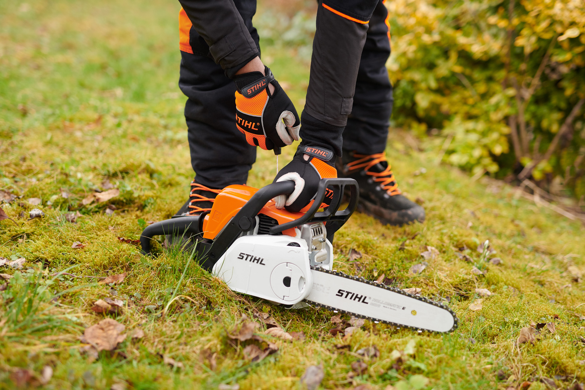 En STIHL MS 180 C-BE motorsav på jorden med en person iført beskyttelseshandsker, der holder håndtaget.