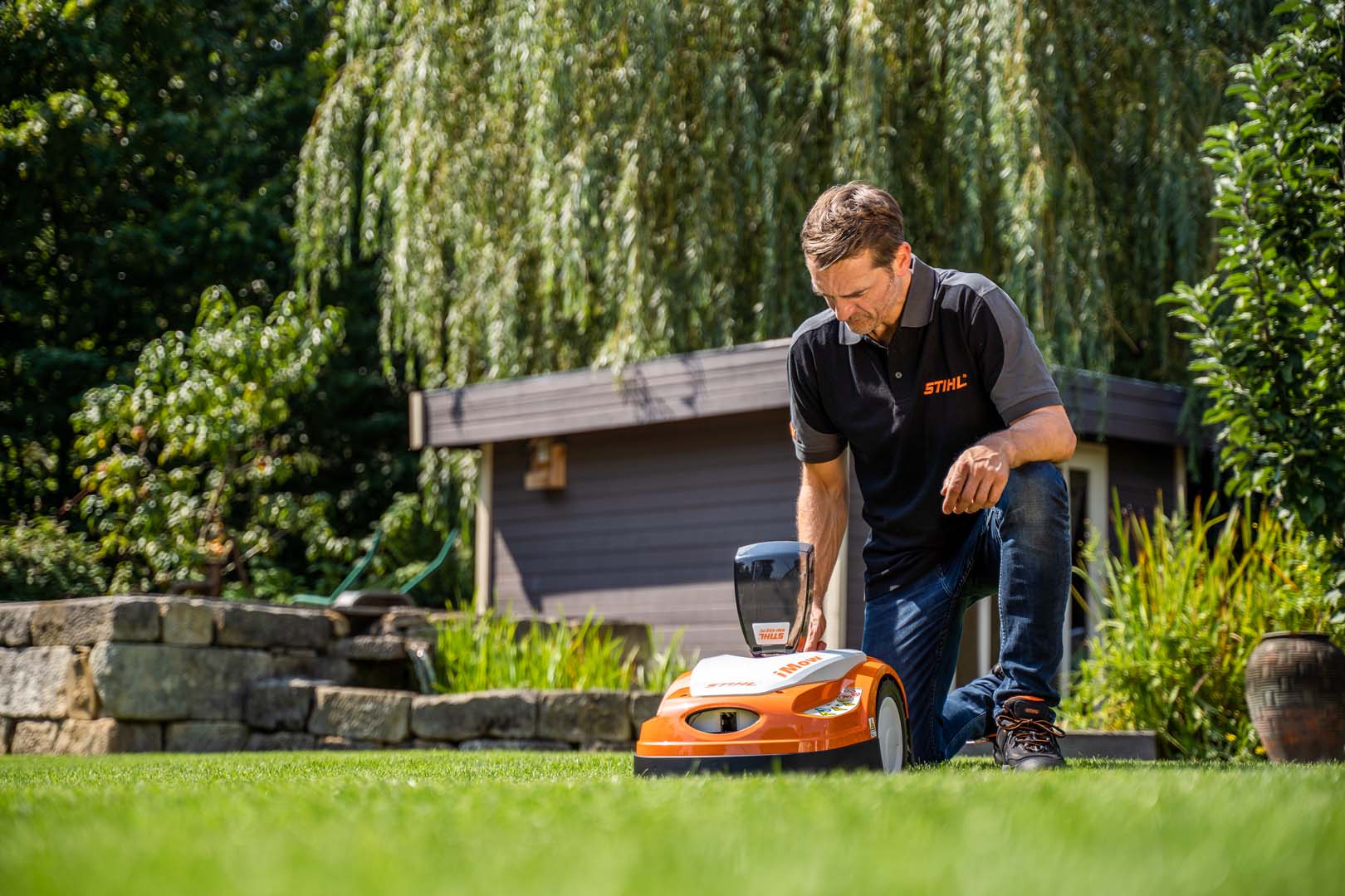 En STIHL-forhandler sidder på knæ for at justere indstillingerne på en STIHL RMI 422 iMOW® robotplæneklipper i en have
