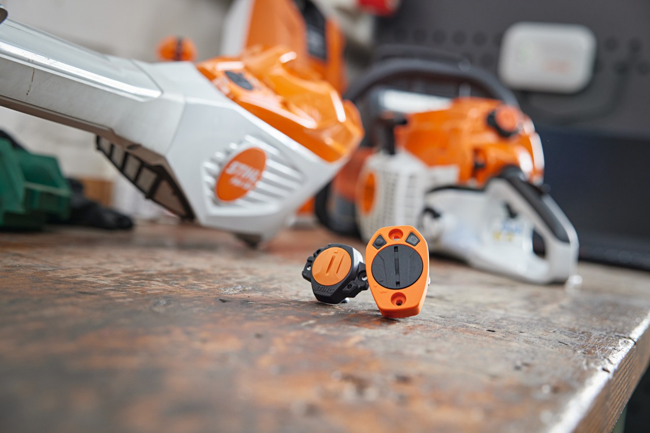En STIHL Smart Connector og en STIHL Smart Connector 2 A på et arbejdsbord. Forskellige STIHL-redskaber kan ses ude af fokus i baggrunden.