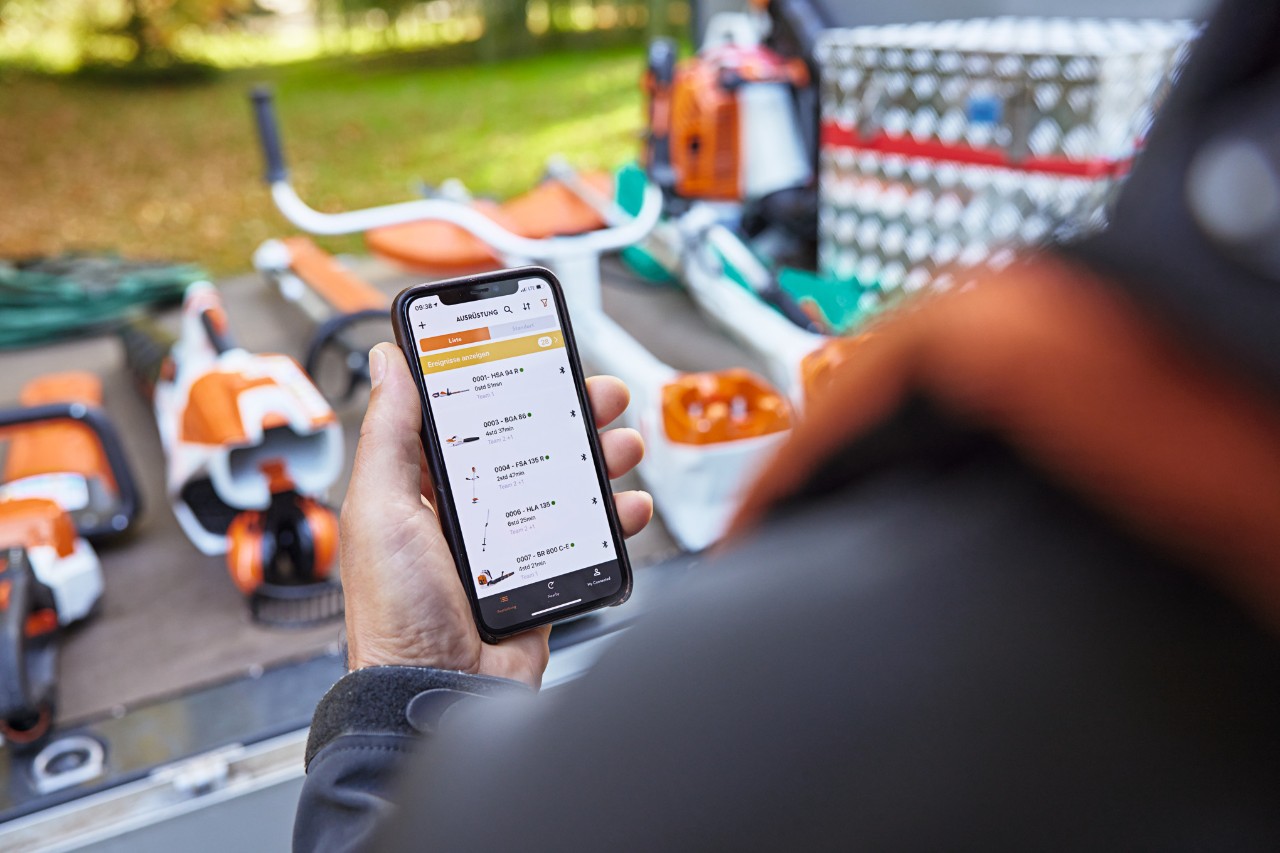 En person, der står foran læsseområdet på en ladvogn, hvorpå der er lagt forskellige STIHL-redskaber. Personen holder en smartphone i hånden med STIHL connected-appen åben. 