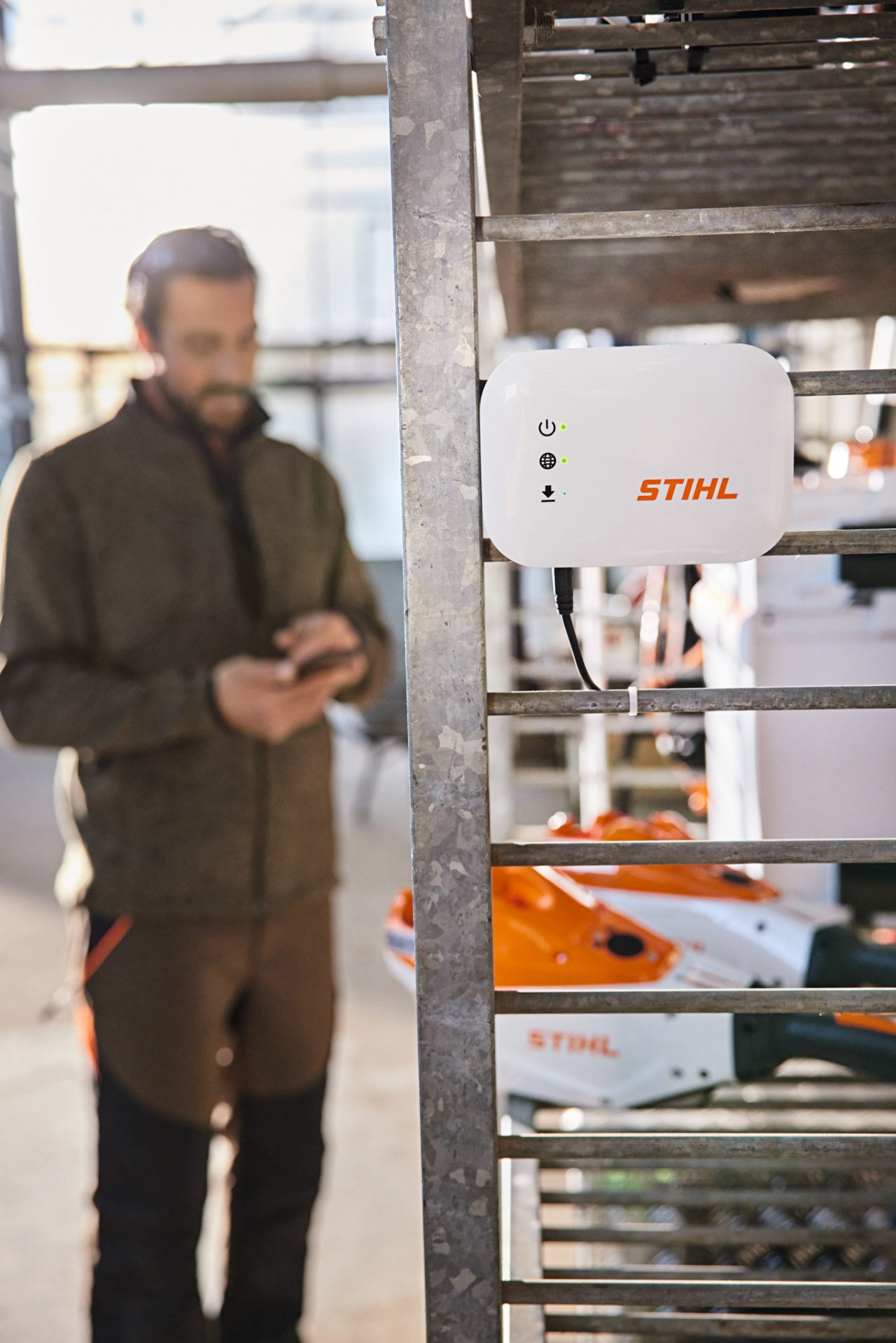 En STIHL connected Box hænger på en værkstedshylde. I baggrunden er der en have- og landskabsarkitekt, der interagerer med sin smartphone. 