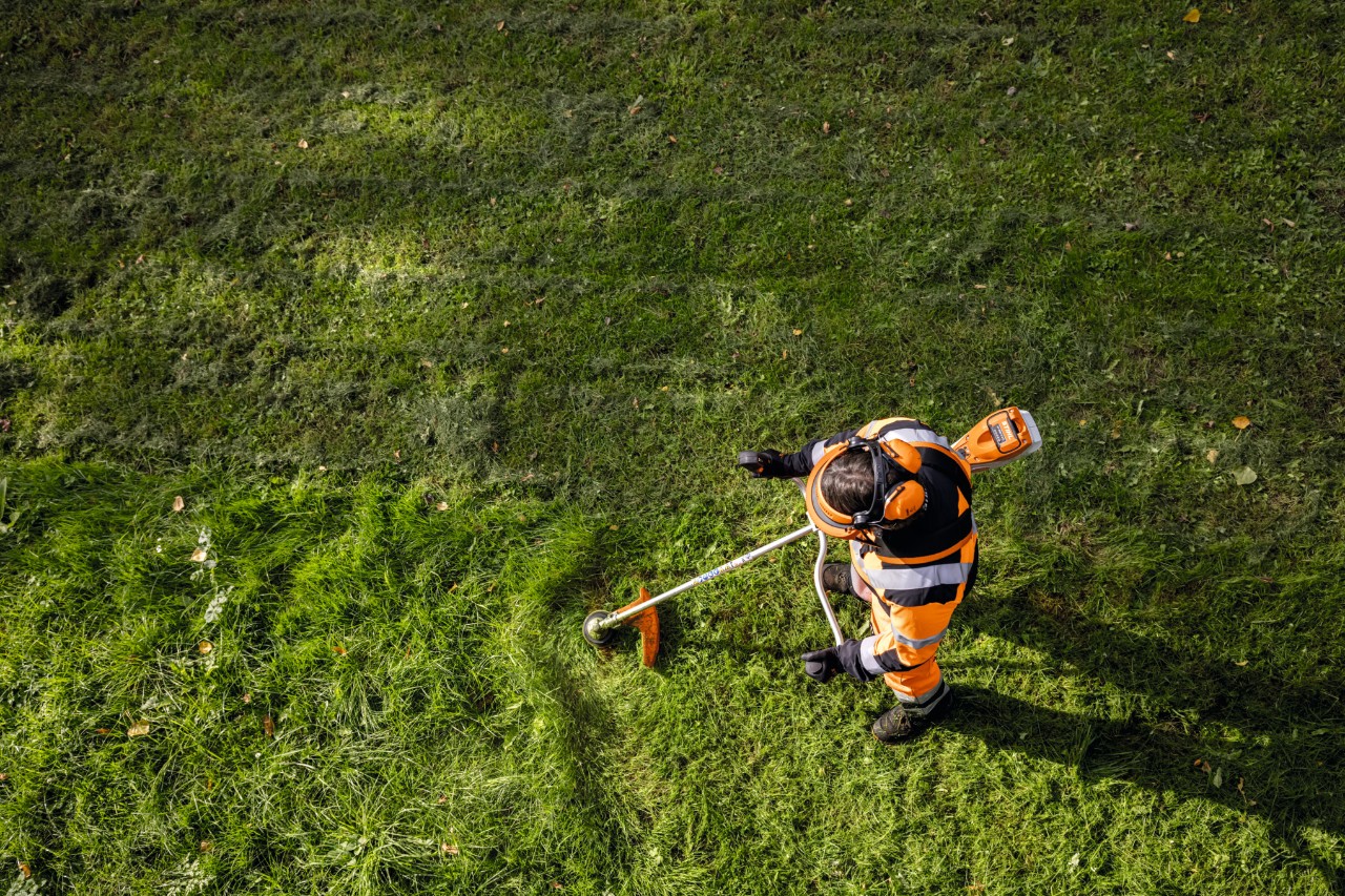 Have- og landskabsarkitekt i orange arbejdstøj klipper højt græs med en STIHL FSA 135 batteridrevet buskrydder