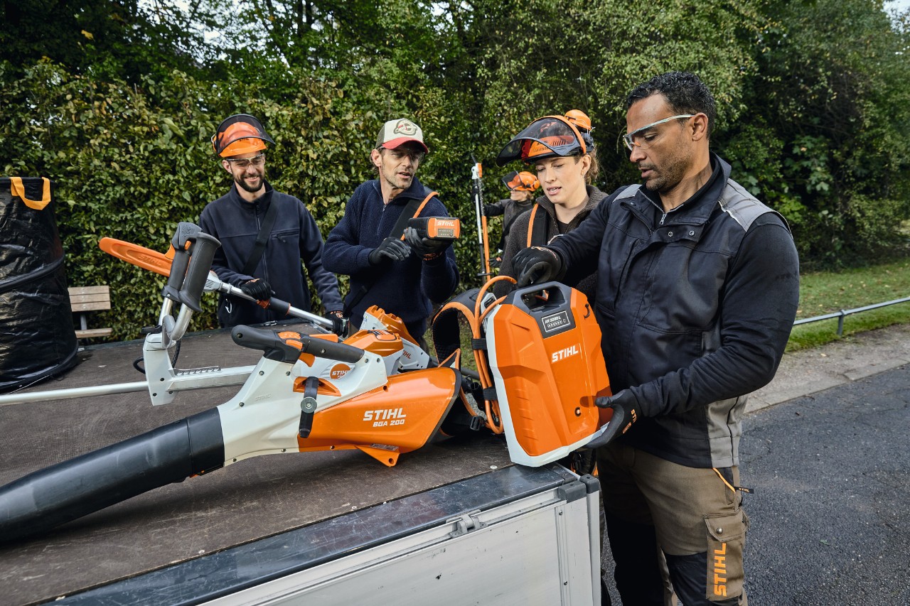 Tre mænd og en kvinde iført STIHL beskyttelsesudstyr står på et køretøjslad med STIHL-elværktøjer, såsom den batteridrevne løvblæser STIHL BGA 200 og rygsækbatteriet STIHL AR 2000 L 