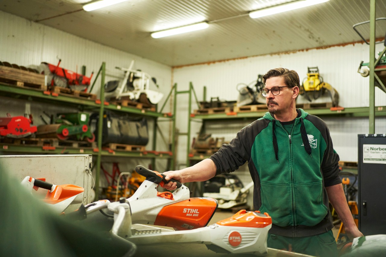 En mand står på et værksted bag en arbejdsflade med STIHL batteriværktøj og rækker ud efter en STIHL batteridrevet løvblæser BGA 200