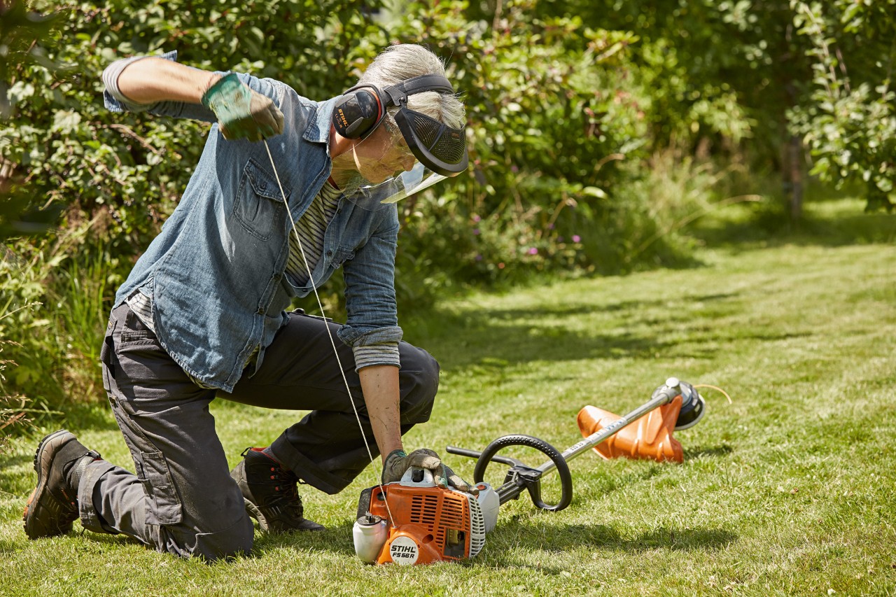 Kvinde med høreværn og ansigtsværn starter en STIHL FS 56 R benzindrevet buskrydder på et græsareal