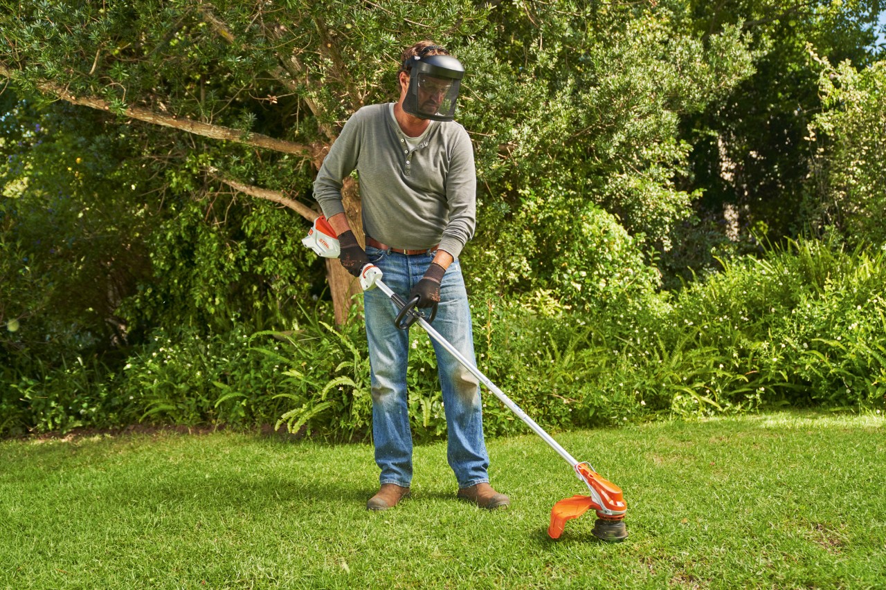 En mand med ansigtsværn klipper græs i en have med den batteridrevne buskrydder STIHL FSA 60