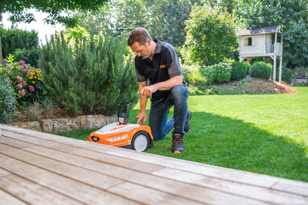 En mand knæler på en græsplæne ved siden af en STIHL iMOW® robotplæneklipper i en have med buske og terrasse