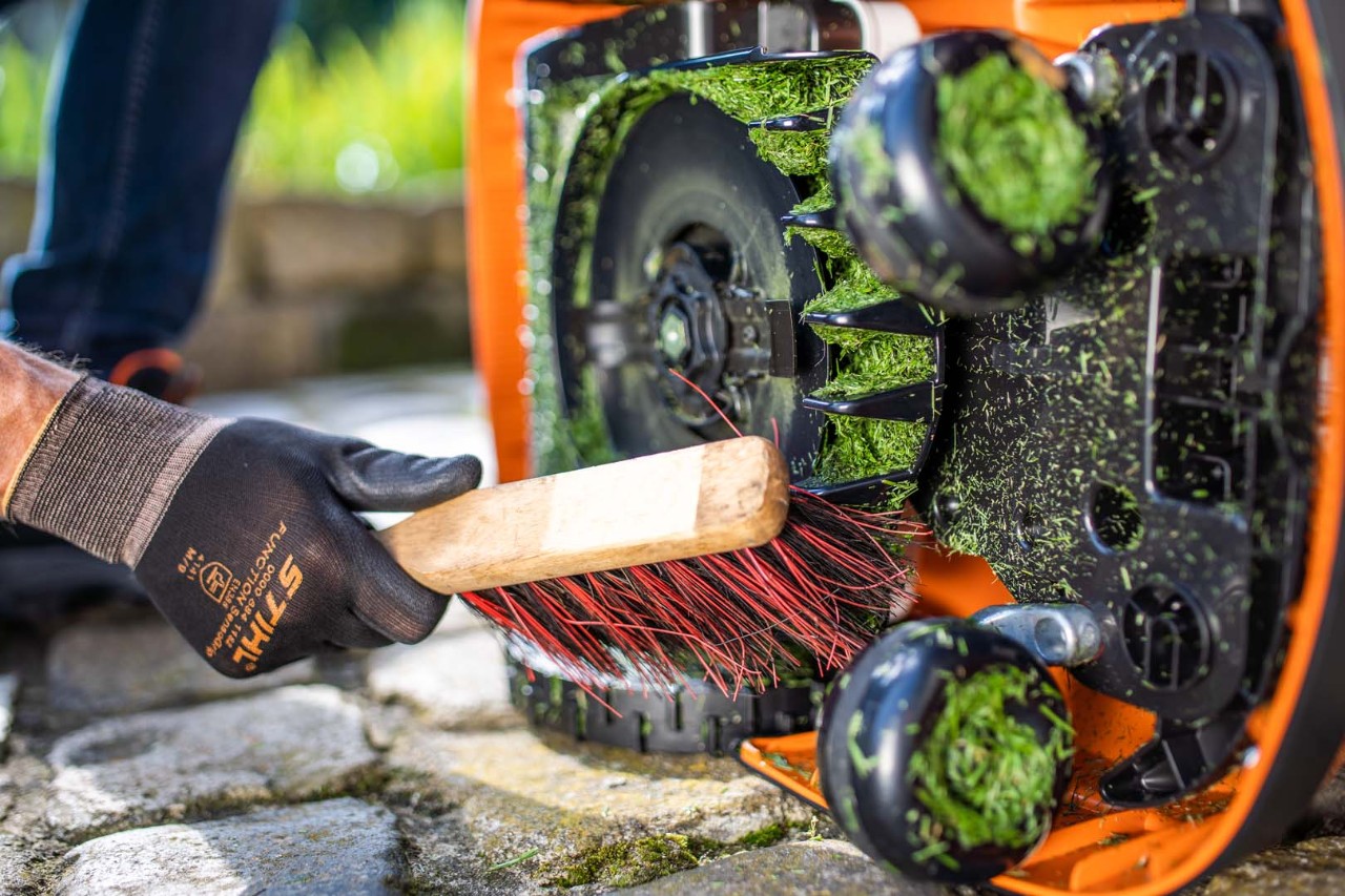 Det græsbelagte dæksel på en STIHL iMOW® robotplæneklipper, der ligger på siden, rengøres med en børste af en person iført handsker