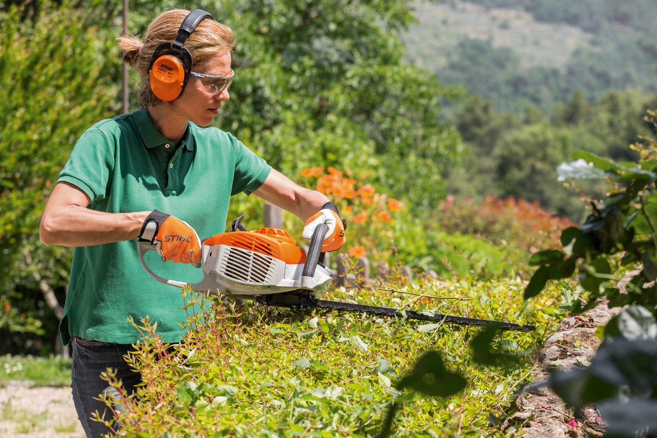 En kvinde med høreværn og beskyttelsesbriller klipper hæk med en STIHL benzinhækkeklipper HS 46