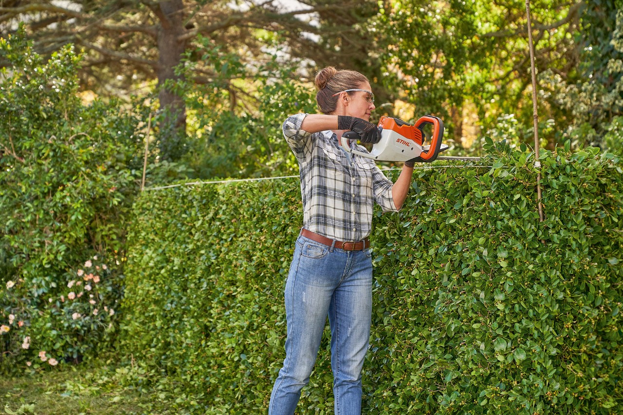 En kvinde med beskyttelsesudstyr udfører hækklipning med STIHL HSA 56 batterihækkeklipper