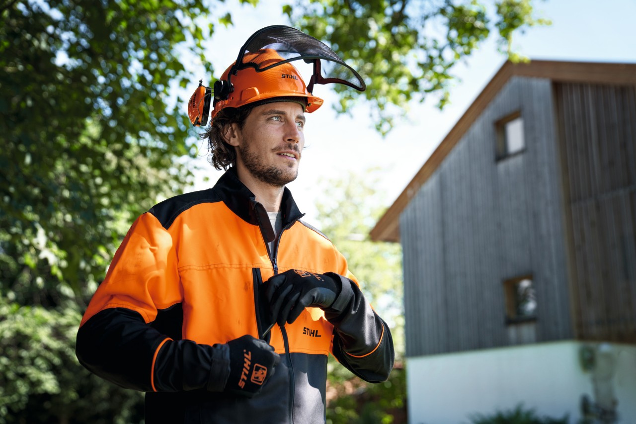 En mand iført orange og sort STIHL-beskyttelsesudstyr, herunder hjelm og handsker, kigger selvsikkert ud af billedet med et hus og træer i baggrunden. 