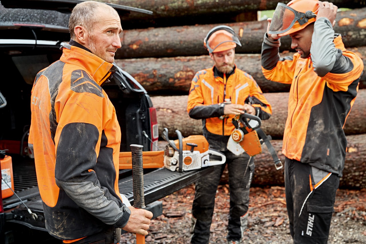Tre mænd i STIHL-beskyttelsestøj gør sig klar til at bruge en STIHL-motorsav foran en træstak.