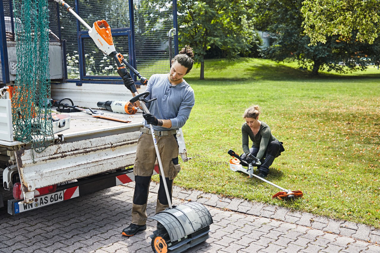 En mand og en kvinde håndterer STIHL-batterimaskiner ved siden af et køretøj med åbent ladeareal, en eng i baggrunden