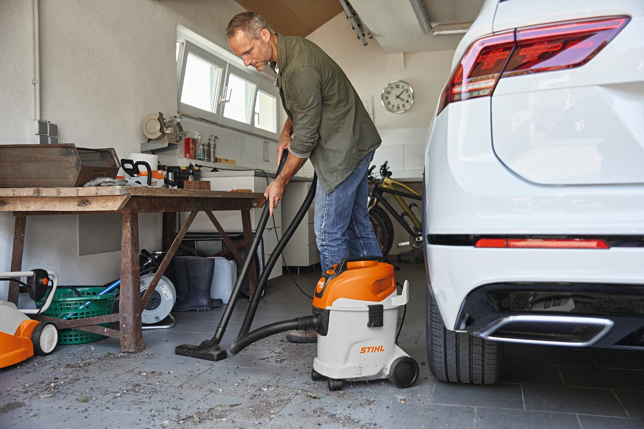 En mand støvsuger gulvet i en garage med en STIHL våd-/tørsuger, ved siden af ham en arbejdsbænk og en hvid bil