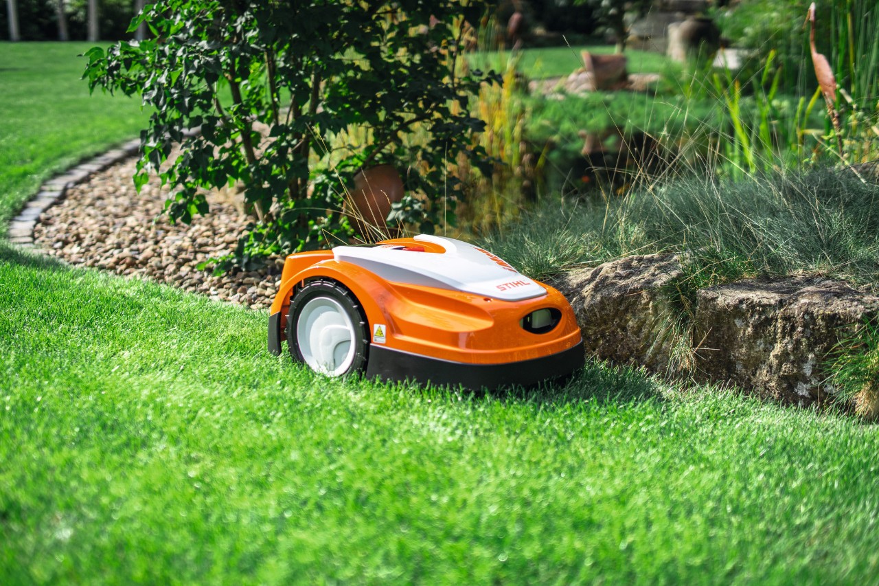 En STIHL iMOW RMI 422 klipper et græsareal i en have ved kanten af en lille stenmur.