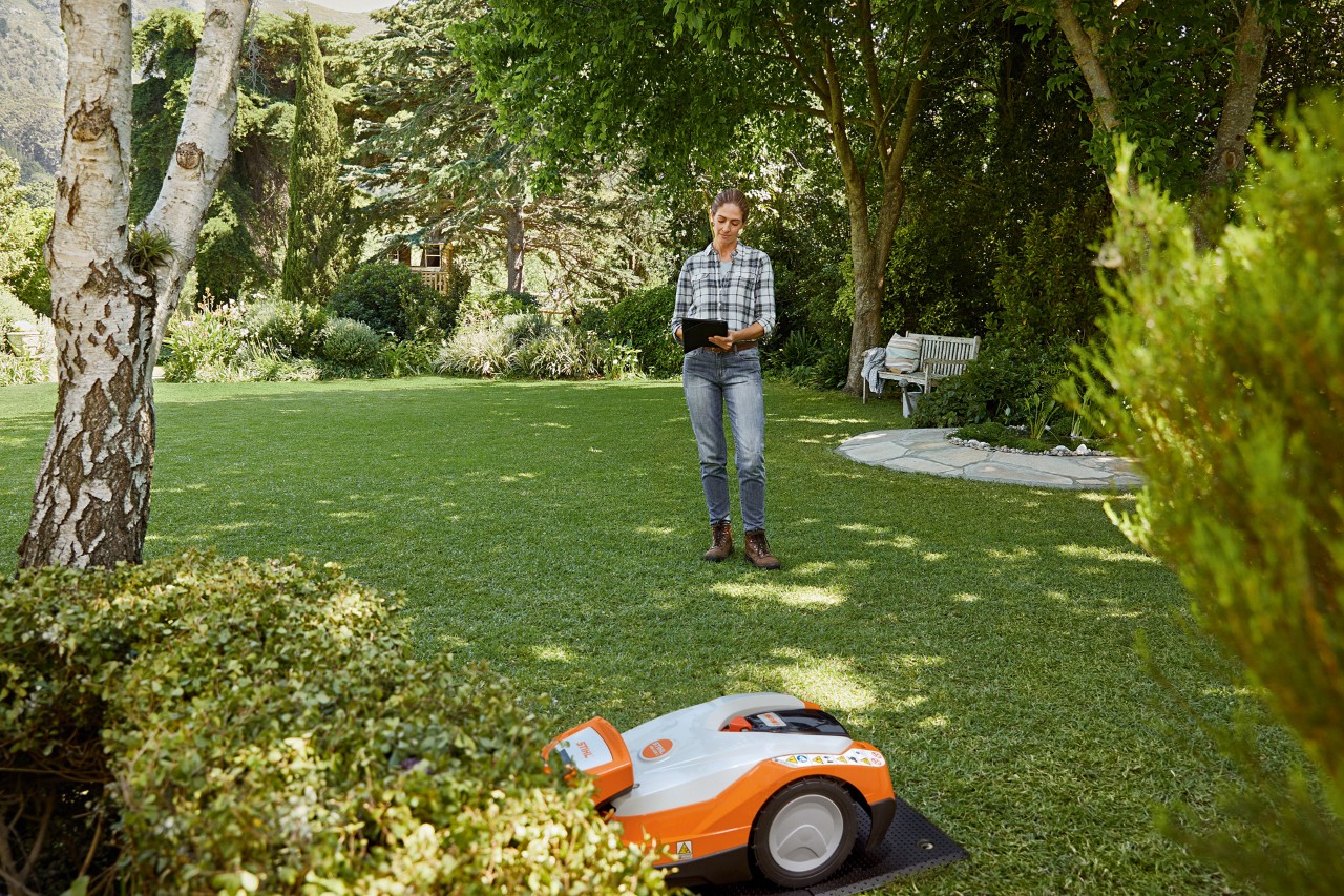 En kvinde bruger en tablet i haven og betjener en STIHL iMOW® 522 robotplæneklipper